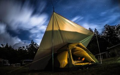 Tente éclairée de l'intérieur la nuit entourée d'arbres et ciel nuageux