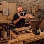 Homme portant masque, ponçant bois sur table dans un atelier rempli d'outils