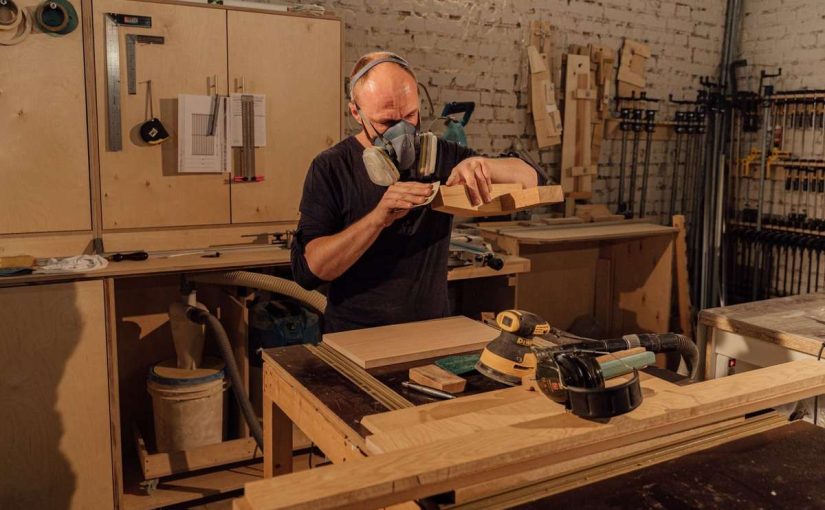 Homme portant masque, ponçant bois sur table dans un atelier rempli d'outils